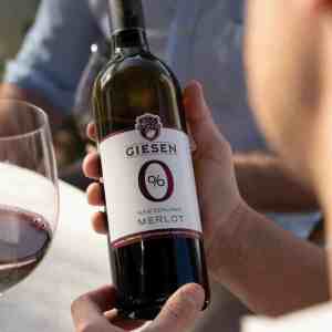 Person holding a bottle of Giesen 0% Merlot New Zealand alcohol free red wine at a table with a glass poured beside it.